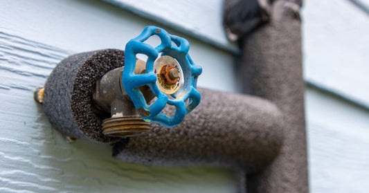 Outdoor water spigot with a blue shutoff valve and insulated pipe, showing signs of rust and weather exposure on a home's exterior wall.