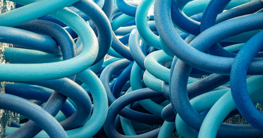 Intertwined loops of thick blue and turquoise pool noodles overlap in a dense, tangled arrangement outdoors.